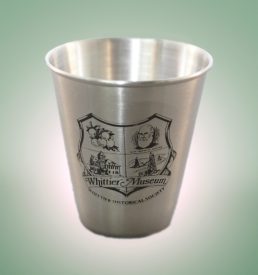 Photograph of stainless steel shot glass with the Whittier Historical Society coat of arms printed in black.