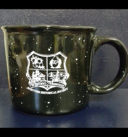 Photograph of coffee mug with the Whittier Historical Society coat of arms printed in white on the mug. Background of mug is black with white speckles in imitation of an enamelware mug.