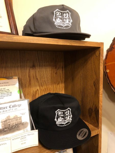 Photograph of two truckers caps with mesh back in black and grey color. Both caps have the Whittier Historical Society coat of arms printed on the front of the cap.