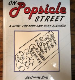 Photograph of the cover of the book On Popsicle Street by Tommy Long. Other text on cover "A book for kids and baby boomers" The drawing on the cover is a child-like drawing of Popsicle street.