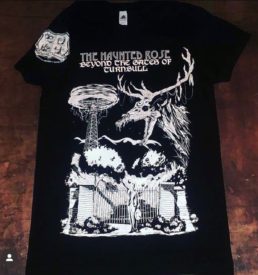 Photograph of 2022 Haunted Rose t-shirt. Shirt is short-sleeved, black color with white graphic on front of shirt showing the electrodome and the gates of hell in Turnbull Canyon. Whittier Historical Society coat of arms on the sleeve. Text "The Haunted Rose Beyond the Gates of Turnbull"