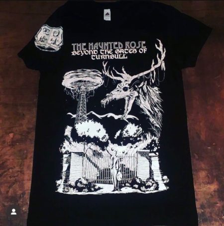 Photograph of 2022 Haunted Rose t-shirt. Shirt is short-sleeved, black color with white graphic on front of shirt showing the electrodome and the gates of hell in Turnbull Canyon. Whittier Historical Society coat of arms on the sleeve. Text "The Haunted Rose Beyond the Gates of Turnbull"